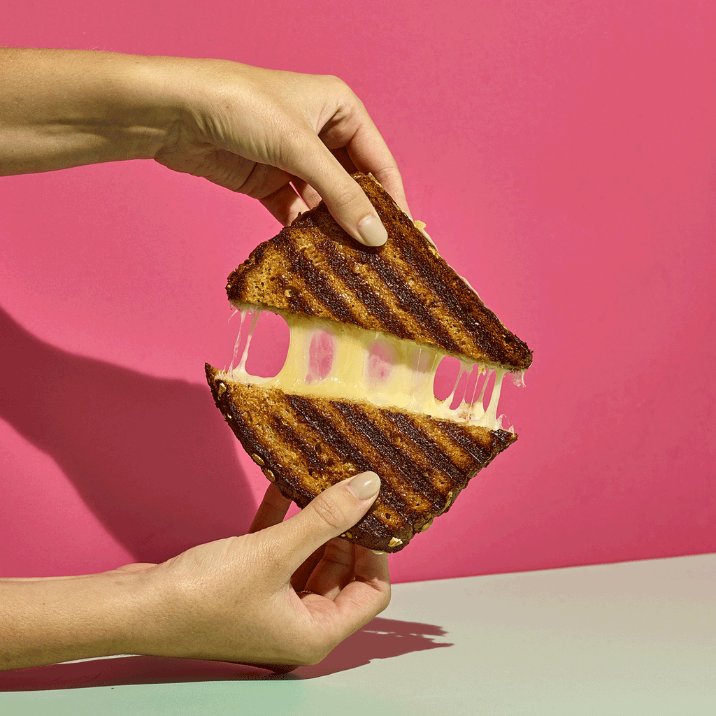 an arm holding a grilled cheese and pulling it apart on a pink and blue background