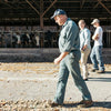 Three people walk past a barn with cows inside. The person in front wears a blue uniform and cap, followed by a smiling individual and another person in a cap. Sunlight casts shadows on the ground covered with dry leaves.