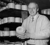 A person stands in a cheese cellar, holding a wheel of cheese labeled Colston Bassett Stilton Cheese. Shelves around them are lined with similar cheeses. The person smiles and wears a light-colored jacket over a sweater and tie.