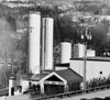 Black and white image of a factory with tall storage tanks labeled Cabot. A building marked Visitor Center is in the foreground. Trees and hills are visible in the background.
