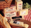 Assorted blocks and packages of Cabot Sharp Vermont Cheddar cheese on a wooden board. A sliced baguette and a bunch of green grapes are nearby on a red and white checkered cloth. A wicker basket is in the background.