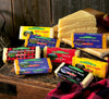 An assortment of Cabot cheese blocks displayed on a wooden surface. Varieties include Seriously Sharp, Extra Sharp, and 50% Light. Some slices are stacked in the background, and a red checkered cloth lies nearby.