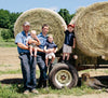 Two men wearing blue shirts stand beside large hay bales on a trailer. One man holds a baby, and the other holds a toddler. A young child stands nearby, raising one arm. A yellow Labrador dog lies on the ground in front. Trees and sky are in the background.
