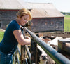 A woman with short blond hair leans over a metal fence, smiling at cows in a pen. She is wearing a blue shirt and jeans. In the background, there are old wooden barns under a clear blue sky. The setting is a rural farm.
