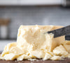 A block of creamy butter with a rough texture is shown on a wooden surface. A knife is partially inserted into the butter, and small pieces are scattered around. The background is softly blurred, giving a kitchen setting impression.