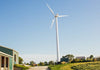 A tall wind turbine stands against a clear blue sky on a sunny day. It is situated near a small building and surrounded by grassy areas and trees. The scene is peaceful and bright.