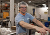 A factory worker wearing a hairnet, safety glasses, and a light blue shirt smiles while operating machinery. He has a visible tattoo on his arm. The background shows industrial equipment and supplies.