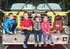 Four children sit smiling on the front bumper of a fire truck labeled New London. They are dressed casually, with one child holding a firefighters hat. A red plaid bag is placed beside them.
