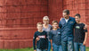 A family of five stands outside in front of a red wooden structure. The parents are smiling, with the father placing an arm around one of the three young boys. Everyone is casually dressed, and the boys are wearing jeans and T-shirts.