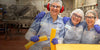 Three workers in hairnets, goggles, and aprons smile and pose inside a food processing facility. They are wearing ear protection and gloves, holding blocks of cheese. Machinery and equipment are visible in the background.