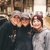 Three smiling people in casual clothing and hats stand close together in a barn. They are surrounded by cows and farm equipment, creating a warm and rustic atmosphere.