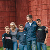 A family of five stands smiling in front of two large, red silo structures. The group includes two adults and three boys, all casually dressed. They seem to be enjoying a pleasant moment together outdoors.