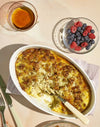 Casserole dish with a portion removed, showing a baked egg and sausage mixture. Next to it is a bowl of fresh raspberries and blueberries, and a glass of amber-colored liquid. Silverware is placed nearby on a napkin.