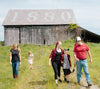 A family of five walks in a grassy area in front of an old wooden barn with 1880 on the roof. Two adults and three children, all casually dressed, are smiling and talking, enjoying the sunny day.