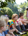 A group of children sit on the grass under a tree on a sunny day. One girl wears a tie-dye shirt and gray boots. They are engaged and looking in the same direction. Trees and a clear sky are in the background.