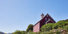 A red barn with a small tower sits on a grassy hillside under a clear blue sky. Green bushes and trees surround the area, with distant hills visible on the left.