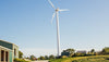 A large wind turbine stands tall against a clear blue sky. It is situated on a grassy landscape with a few small buildings and trees nearby.