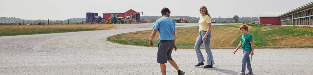A man, woman, and child walk on a gravel path near barns and a tractor on a farm, with open fields and a clear sky in the background.