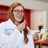 A woman sampling a cube of cheese, she's in a white lab coat and she's a Cheese Grader