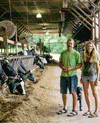 A man and a woman stand in a barn with cows. The man is wearing a green shirt and shorts, and the woman is in a gray top and shorts. A black dog stands behind them. The barn has fans on the ceiling, and cows are eating from a feeding area.