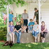 A group of people and a dog sit and stand on a porch in front of a white house. There is a U.S. flag hanging on the left. The group includes men and women, casually dressed, and a brown dog lies on the grass. Trees are in the background.