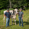 A man and woman stand beside a black and white cow in a grassy field. The man is wearing a black t-shirt and cap, while the woman holds the cows rope and wears a dark t-shirt. Other cows graze in the background near trees.