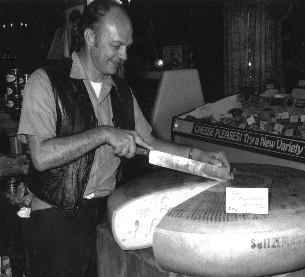 A man wearing a leather vest slices a large round cheese wheel in a store. A sign on the cheese reads Switzerland, and theres a display in the background with a sign saying Cheese Please! Try a New Variety.