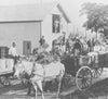 A vintage black and white image of workers loading barrels onto horse-drawn wagons outside a wooden building. A man stands in the doorway above, guiding the operation. Horses are hitched to wagons filled with barrels. Trees and equipment are visible.