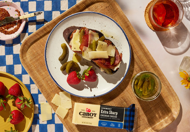 a tartine with cabot cheddar, meats, pickles and strawberries on the side with a blue and white checkered table cloth.