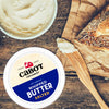 On a wooden table, an open container of Cabot Creamery Whipped Salted butter sits beside a wooden spreader with butter. A piece of sesame-crusted bread is partially visible on brown paper.