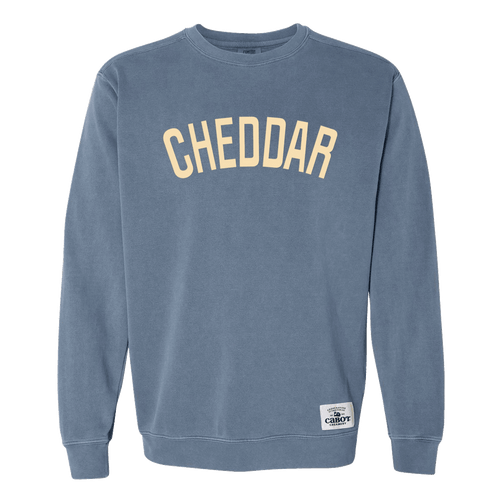 The Cheddar Crewneck by New Duds is a blue sweatshirt featuring the word “CHEDDAR” in large, cream-colored letters across the chest and a small white patch near the hem.