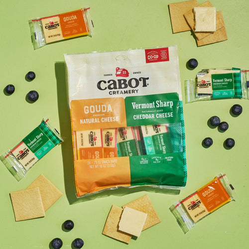A bag of Cabot Creamery Sharp & Gouda Variety Pack, with snack-size Vermont Sharp Cheddar and Gouda cheese bars, sits on a green background surrounded by wrapped cheese sticks, crackers, cheese slices, and blueberries.