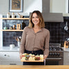 A woman with shoulder-length brown hair smiles in a modern kitchen with a black tile backsplash, holding the Cabot X That Cheese Plate: Ultimate Charcuterie Gift Box by Cabot Creamery, filled with assorted cheeses and grapes.