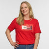 A smiling woman with long blonde hair wears a red Seriously Sharp Cheddar Bar T-shirt by New Duds and blue jeans, standing with one hand on her hip against a white background. She has a Cabot Creamery name tag on her chest.