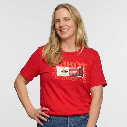 A smiling woman with long blonde hair wears a red Seriously Sharp Cheddar Bar T-shirt by New Duds and blue jeans, standing with one hand on her hip against a white background. She has a Cabot Creamery name tag on her chest.