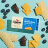 A block of Cabot Creamery Seriously Sharp Yellow Cheddar Cheese sits on a light blue background, surrounded by cheddar slices, graham crackers, chocolate pieces, and blueberries.