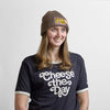 A person wears a Where Life is Cheddar Beanie in olive ribbed-knit with a rectangular patch, paired with a black t-shirt that says cheese the Day.