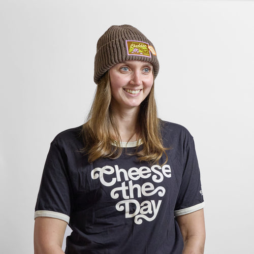 A person wears a Where Life is Cheddar Beanie in olive ribbed-knit with a rectangular patch, paired with a black t-shirt that says cheese the Day.