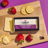 A wooden board features a partially sliced block of Cabot Creamerys Vintage Choice Cheddar Cheese, aged 24 months. It is paired with round crackers and fresh strawberries on a purple surface.