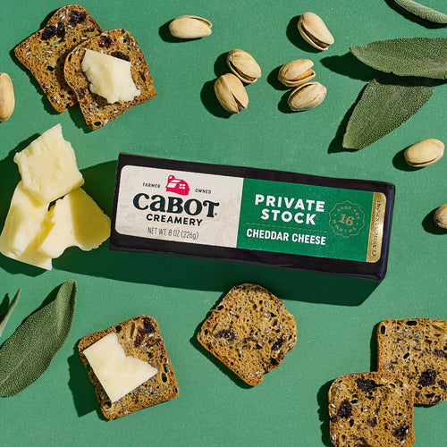 A block of Cabot Creamerys Private Stock Cheddar Cheese, naturally aged for buttery smoothness, sits on a green surface surrounded by multigrain cracker slices topped with cheese, pistachios, and sage leaves.