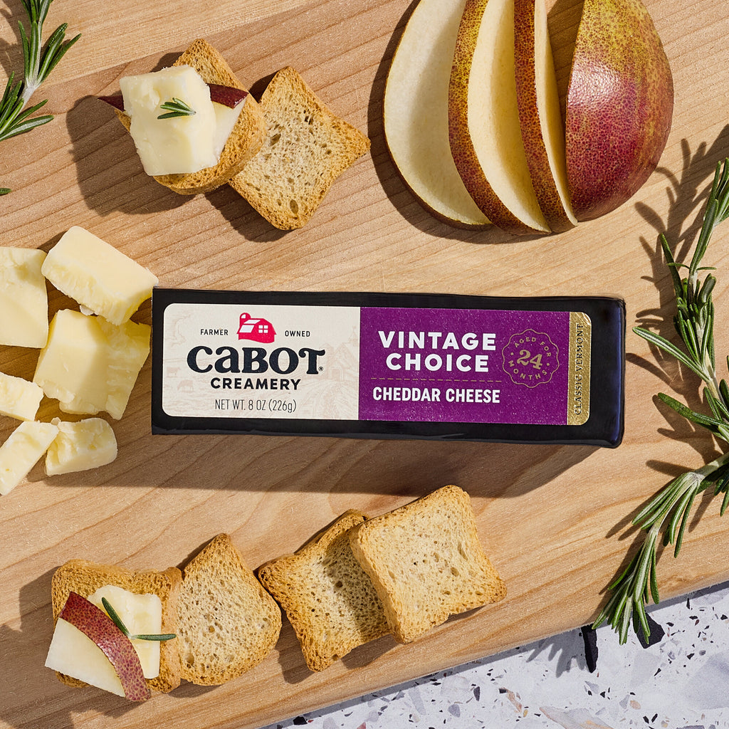 A block of Cabot Creamerys Vintage Choice Cheddar Cheese, aged 24 months, lies on a wooden board surrounded by cheese cubes, crispy bread slices, rosemary sprigs, and sliced pears.