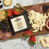 A wooden board features super-premium Cabot Creamery cheddar slices, salami, strawberries, blackberries, and crackers. Aged cheddar from Cabot Creamery shines with its tangy bite. The spread is complemented by a small dish of jam on a marble surface.