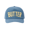 The Butter Corduroy Hat in blue by New Duds features the word “BUTTER” embroidered in bold cream lettering on the front and is finished with a classic snapback closure for an adjustable fit.
