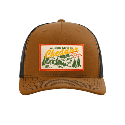 The New Duds Where Life is Cheddar Trucker Hat is a brown and black snapback with an adjustable fit, featuring a patch that reads “Where Life Is Cheddar Vermont USA” along with a rural landscape illustration of trees and a barn.