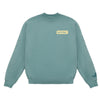A teal Life’s Better With Butter Crewneck by New Duds, featuring a small yellow rectangular patch with “BUTTER” on the left chest and a subtle dark logo near the left wrist.