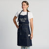 A woman stands smiling against a plain light background, wearing the New Duds “I Aim To Cheese” apron over a white T-shirt.