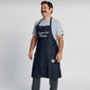 A man stands smiling with his hands by his sides, dressed in a light gray t-shirt and dark pants, wearing a blue denim “I Aim To Cheese” apron by New Duds, against a plain white background.