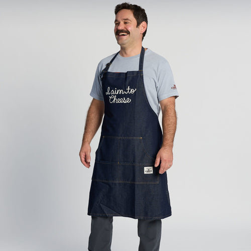 A man stands smiling with his hands by his sides, dressed in a light gray t-shirt and dark pants, wearing a blue denim “I Aim To Cheese” apron by New Duds, against a plain white background.