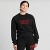A person with dark hair in a bun smiles while wearing the New Duds Seriously Sharp Chainstitch Crewneck—a black sweatshirt featuring “Seriously Sharp” in red script—paired with matching black sweatpants with red text, standing against a plain light background.