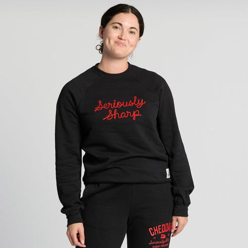 A person with dark hair in a bun smiles while wearing the New Duds Seriously Sharp Chainstitch Crewneck—a black sweatshirt featuring “Seriously Sharp” in red script—paired with matching black sweatpants with red text, standing against a plain light background.